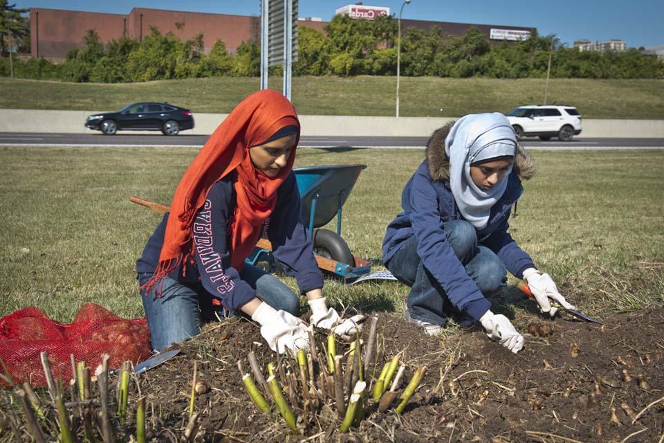 Two 博彩网址大全 students kneel next to a gardening plot, wearing gloves, headscarves and digging in the dirt.